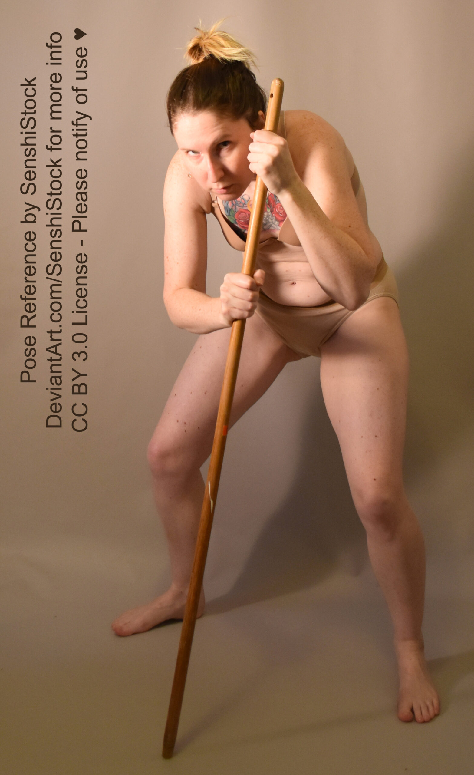 A white woman in a bra and undies crouched over looking like a bridge troll, holding a staff against the ground.