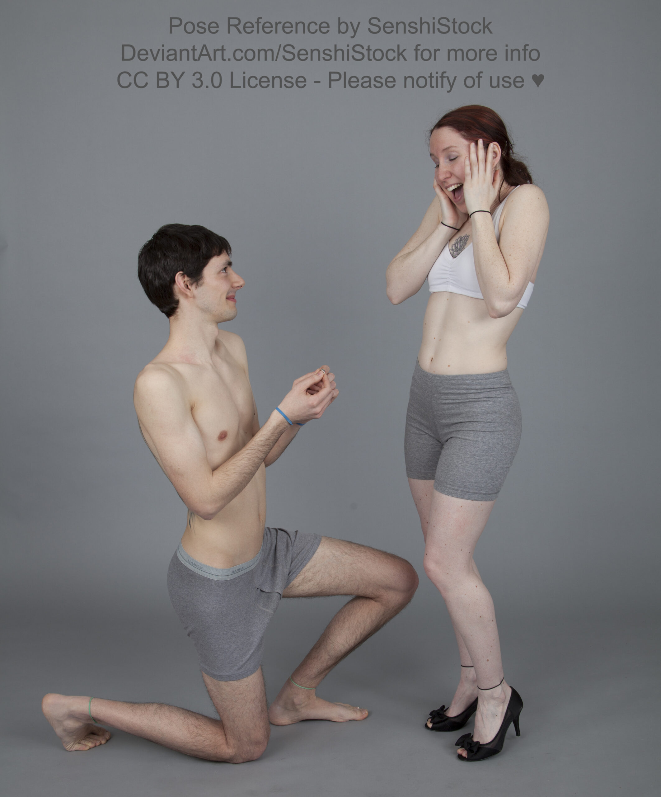 A skinny white man down on one knee, doing a mock proposal to a skinny white woman wearing heels, her hands on her face in shock.
