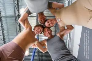 Four white models with their heads together, linking arms with one another, looking down and smiling at the camera. Two models are female and two are male, and they are all wearing grey or neutral skin tone tight clothing.