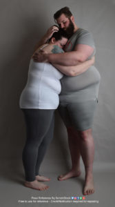 Two fat white models wearing skin tight clothing are wrapped in an embrace in each other's arms, tightly pressed up against each other. The shorter one is pressing their face into the taller one. The shot is in profile.