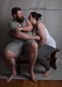 Two fat white models wearing skin tight clothing are sitting on a dark brown bench. The man is sitting forward but leaning back as the woman is leaned forward and attempting to bite his upper arm.