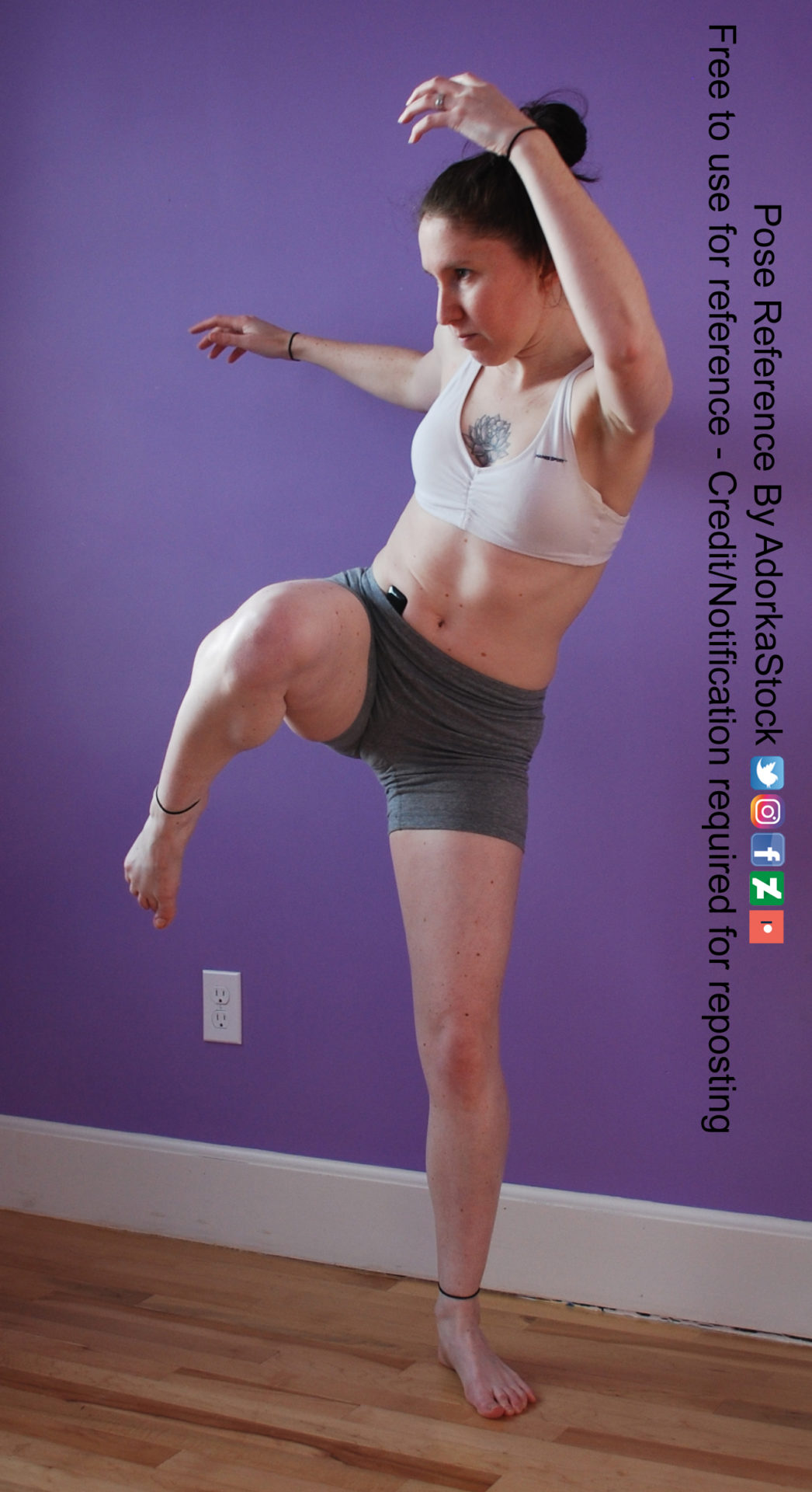 Thin white woman wearing white sports bra and form-fitting grey shorts, standing on one leg while the other is raised upward as if preparing for a kick. Her arms are raised in the air, one in front, bent at the elbow. The other is out-stretched behind her.