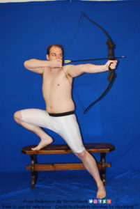 A white man in skin tight shorts, one leg up on a bench, aiming with a bow up towards the right.