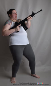 A fat white female model wearing skin tight clothing is standing, legs apart, while holding a gun, pointed up and to the right, with proper trigger finger, looking way too excited to be holding a weapon, as she's leaning back with an open grin on her face.
