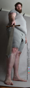 A fat white male model wearing skin tight clothing is seen from an extreme worm's eye view, looking up at him, as he's standing and looking back down at the camera, with a sword in one hand and gestured toward the viewer.
