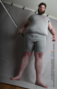 A fat white male model wearing skin tight clothing is seen from an extreme worm's eye view, looking up at him, as he's standing and looking back down at the camera, with a sword in one hand and the other relaxed, feet apart.