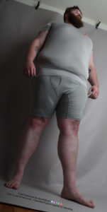 A fat white male model wearing skin tight clothing is seen from an extreme worm's eye view, looking up at him, as he's standing and looking up and away, arms resting at his sides and feet apart.