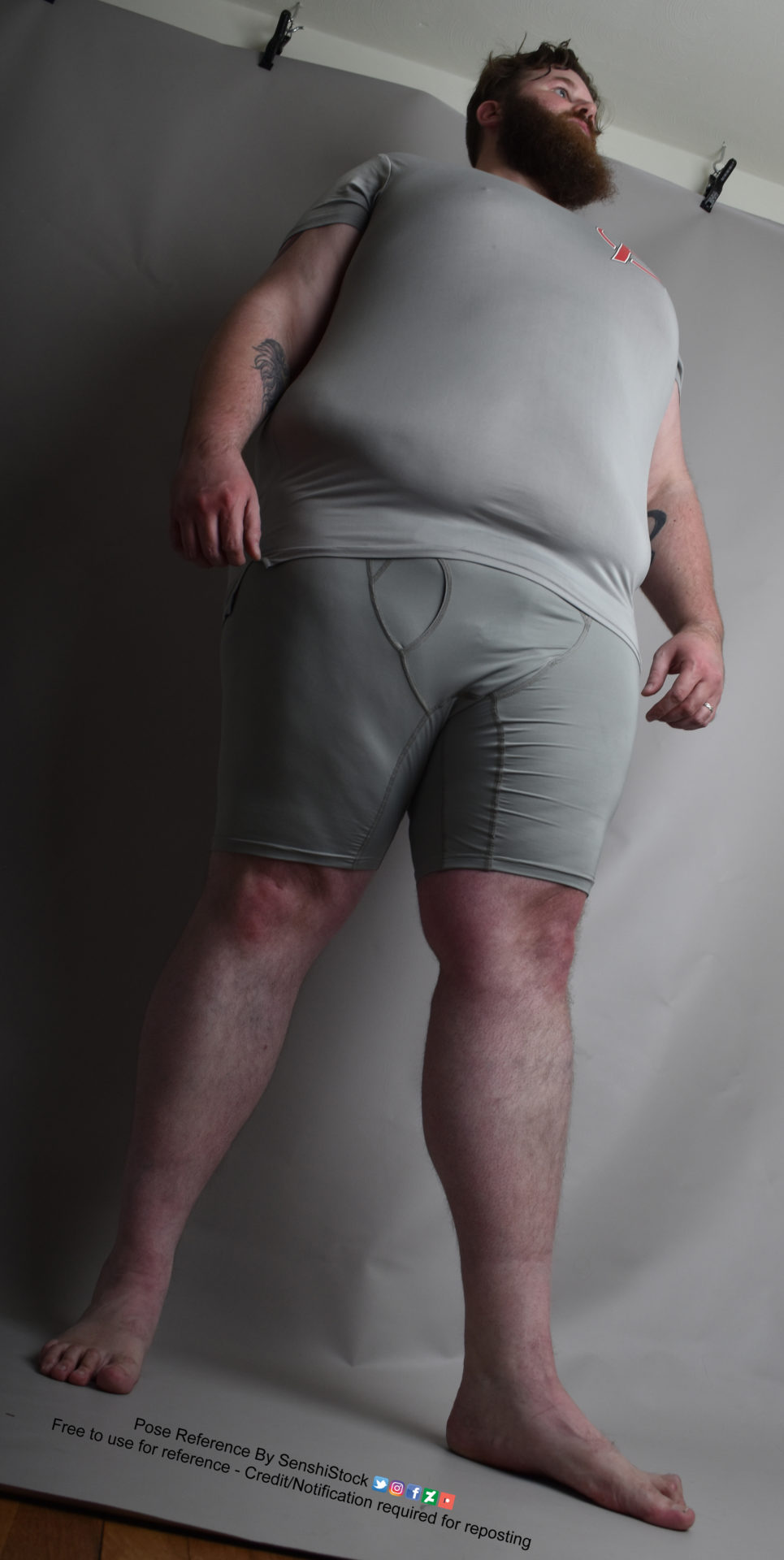 A fat white male model wearing skin tight clothing is seen from an extreme worm's eye view, looking up at him, as he's standing and looking up and away, arms resting at his sides and feet apart.