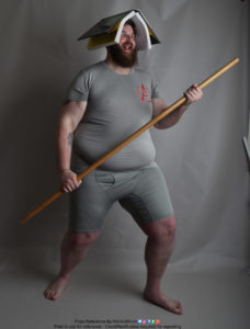 A fat white model wearing skin tight clothing is standing, legs apart in a stable stance, while holding a bo staff with both hands, an open book covering his head like a not super great hat, as he's making a silly face.