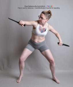 Thin white woman wearing a white sports bra and form-fitting grey shorts, and glasses, ever-so-slightly crouched, legs spread apart, as she's holding a knife in each hand, arms spread outward, her gaze to the right (viewer left).
