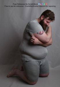 A fat white model wearing skin tight clothing is on his knees, at a 3/4 angle, arms wrapped around himself and head down.