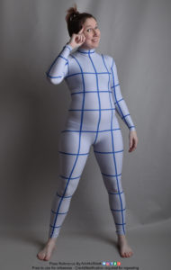 Thin white woman wearing a form-fitting zentai suit (full body outfit with lines to help with perspective), standing facing the camera with one arm relaxed and down, the other raised with a finger pointing at her forehead as she's giving you a big grin.