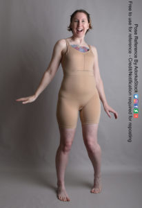 Thin white woman wearing a form-fitting beige body suit, standing with her toes slightly pointed inward, arms down at her side, with a massive grin on her face.