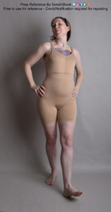 Thin white woman wearing a form-fitting beige body suit, standing with one foot out like she's walking. Her hands are on her hips and her head is slightly turned to the right (viewer left) with a soft smile.