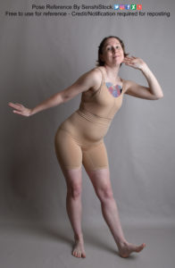 Thin white woman wearing a form-fitting beige body suit, standing while leaning out to the side, one foot out and raised with just the heel touching the ground. One arm is out to the side and the other is raised to her ear.