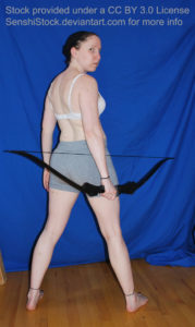 White woman holding a bow in her right hand, standing, looking over her shoulder at the camera.