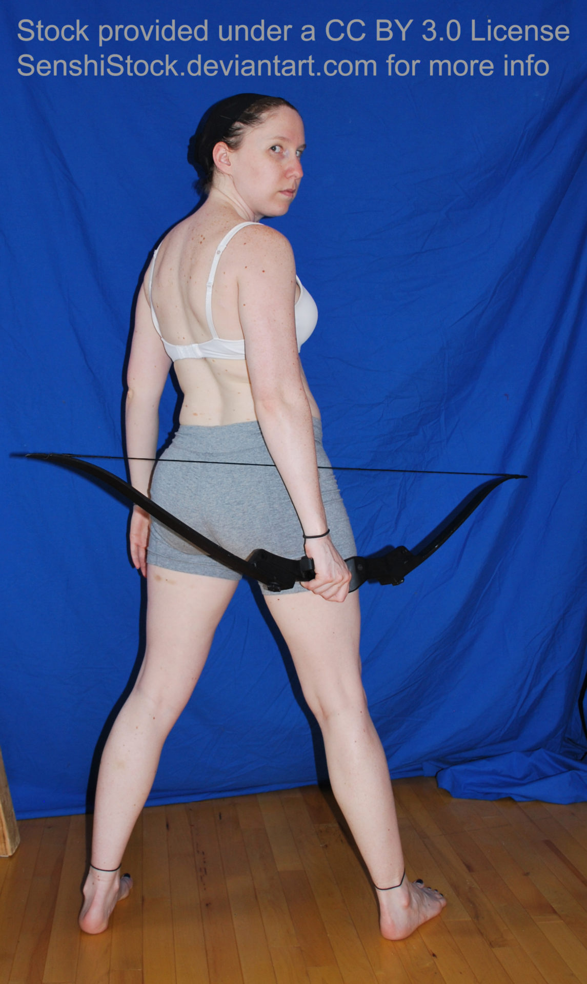 White woman holding a bow in her right hand, standing, looking over her shoulder at the camera.