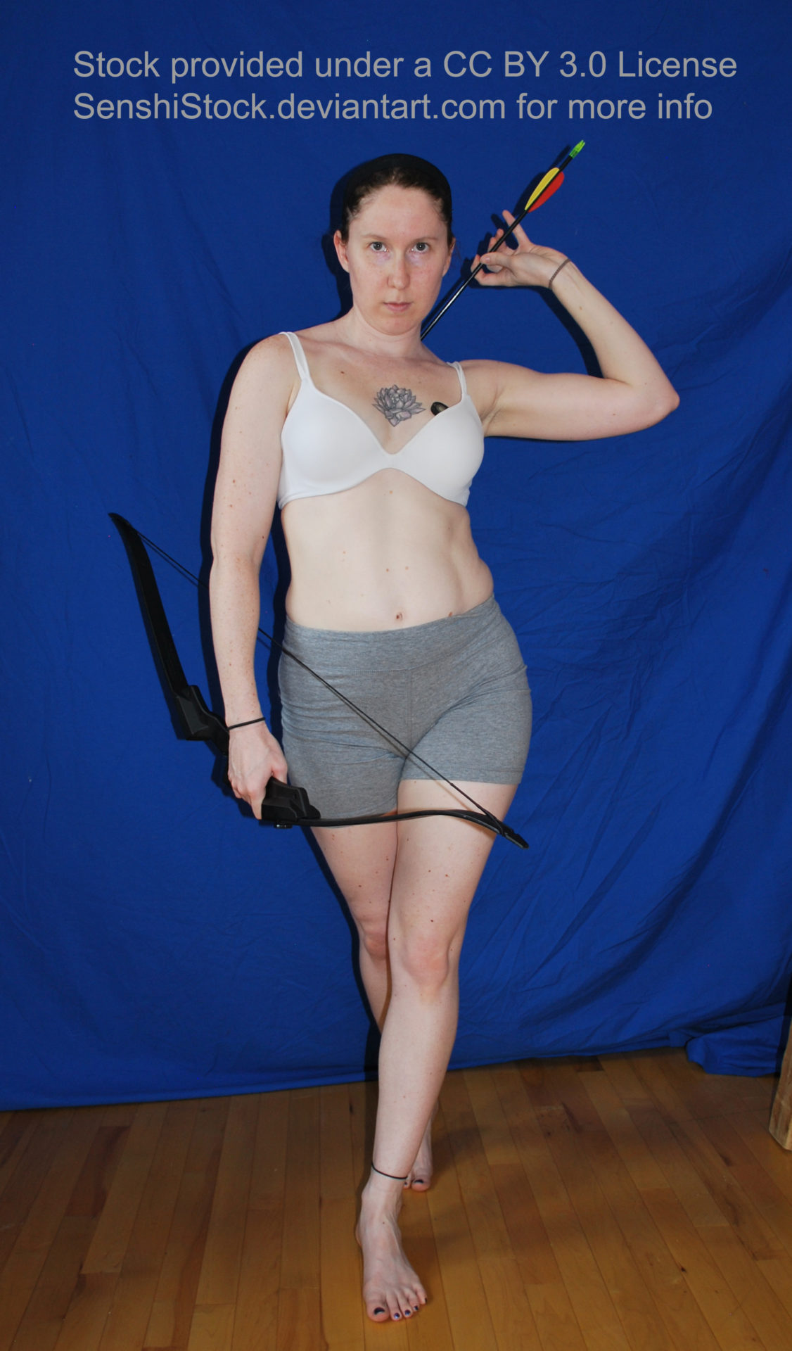 White woman walking forward towards the viewer, one arm over her right shoulder gripping an arrow from her back, her left hand holding a bow down by her thigh. Her facial expression looks determined. She is wearing a white bra and grey bike shorts.