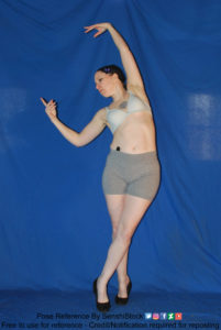 Thin white woman wearing white sports bra and form-fitting grey shorts and black shoes is standing with her body facing the camera and with her legs crossed, as she's leaned slightly to the side, with both arms are raised, one slightly bent at the elbow with her hand curled in, the other is straight above her head, as if she's dancing.