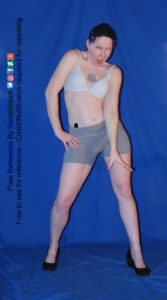 Thin white woman wearing white sports bra and form-fitting grey shorts and black shoes is standing facing the camera with her legs apart, hip lifted to one side, with one hand resting at the top of her thigh, the other arm is straight down, fingers spread apart. She's winking with an open mouth.