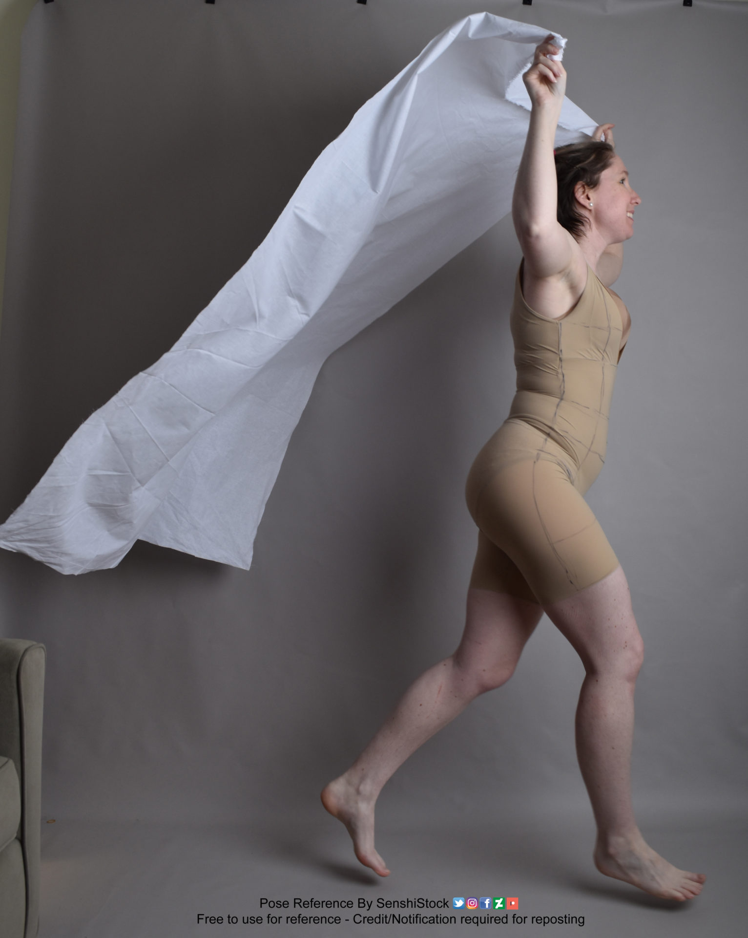 Thin white woman wearing a form-fitting beige body suit, standing in profile, walking to the right, with her arms raised overhead as she holds on to a giant white sheet trailing behind her.