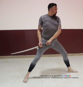 White man standing in a gymnasium wearing a grey guit holding a katana down by his thigh.