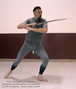 A white man standing with his front to the camera, wearing a grey gym suit, a katana in one hand angled over his shoulder, his other bent in front of him.