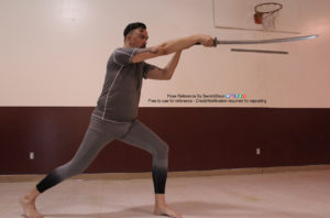 A man in a grey gym suit standing, knees bent, pointing a katana out to the right.