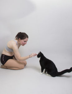 A thin, white model wearing a sports bra and shorts, sitting on the floor on her knees, leaning forward and is giving a gentle scratch to the forehead of a cat that's sitting next to her.