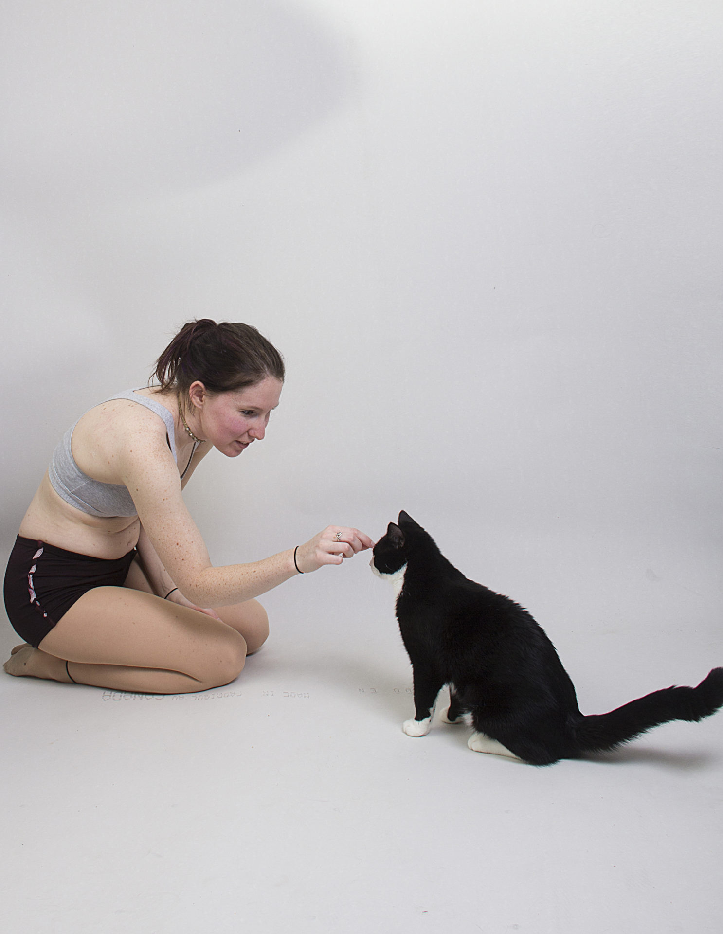 A thin, white model wearing a sports bra and shorts, sitting on the floor on her knees, leaning forward and is giving a gentle scratch to the forehead of a cat that's sitting next to her.