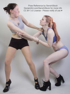 Two thin, white, female models wearing sports bras and short shorts with black high heels, in a scuffle. One is trying to push the other away, face scrunched up and angry. The other is crouched and looking up at the first, desperately trying to hold on.