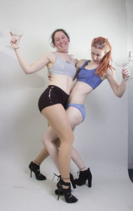 Two thin, white, female models wearing sports bras and short shorts with black high heels, are drunk dancing, margarita glasses in hand. They're both holding on to each other as they look like they are struggling to stand up but also dance together at the same time.
