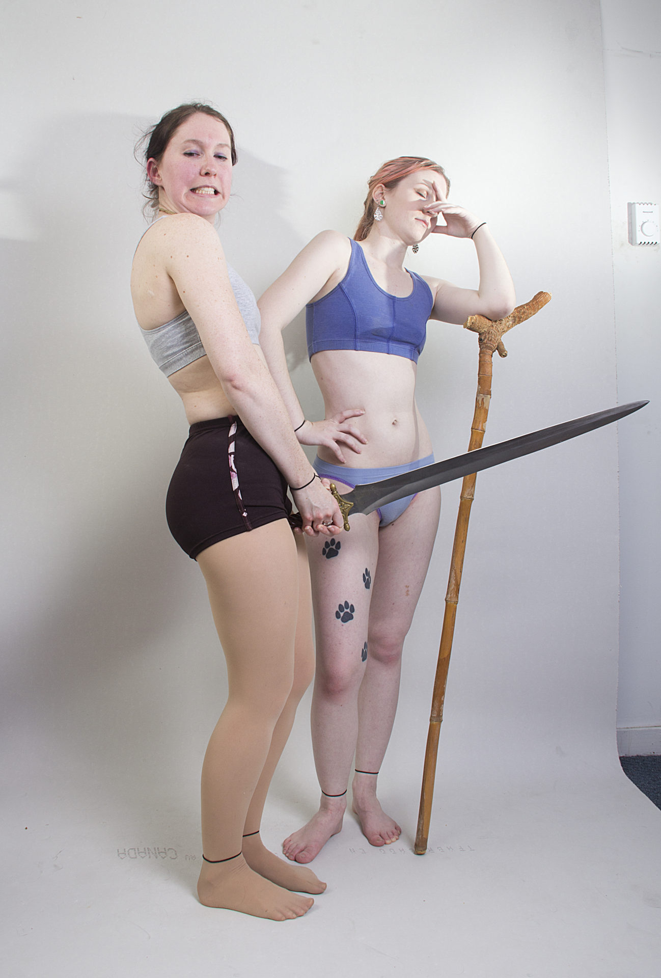 Two thin, white, female models wearing sports bras and short shorts, with one standing leaning on a wooden staff, clearly exasperated with the other woman who is standing profile, holding up a massive sword that is definitely not a phallic symbol, making a face of drunken pride.