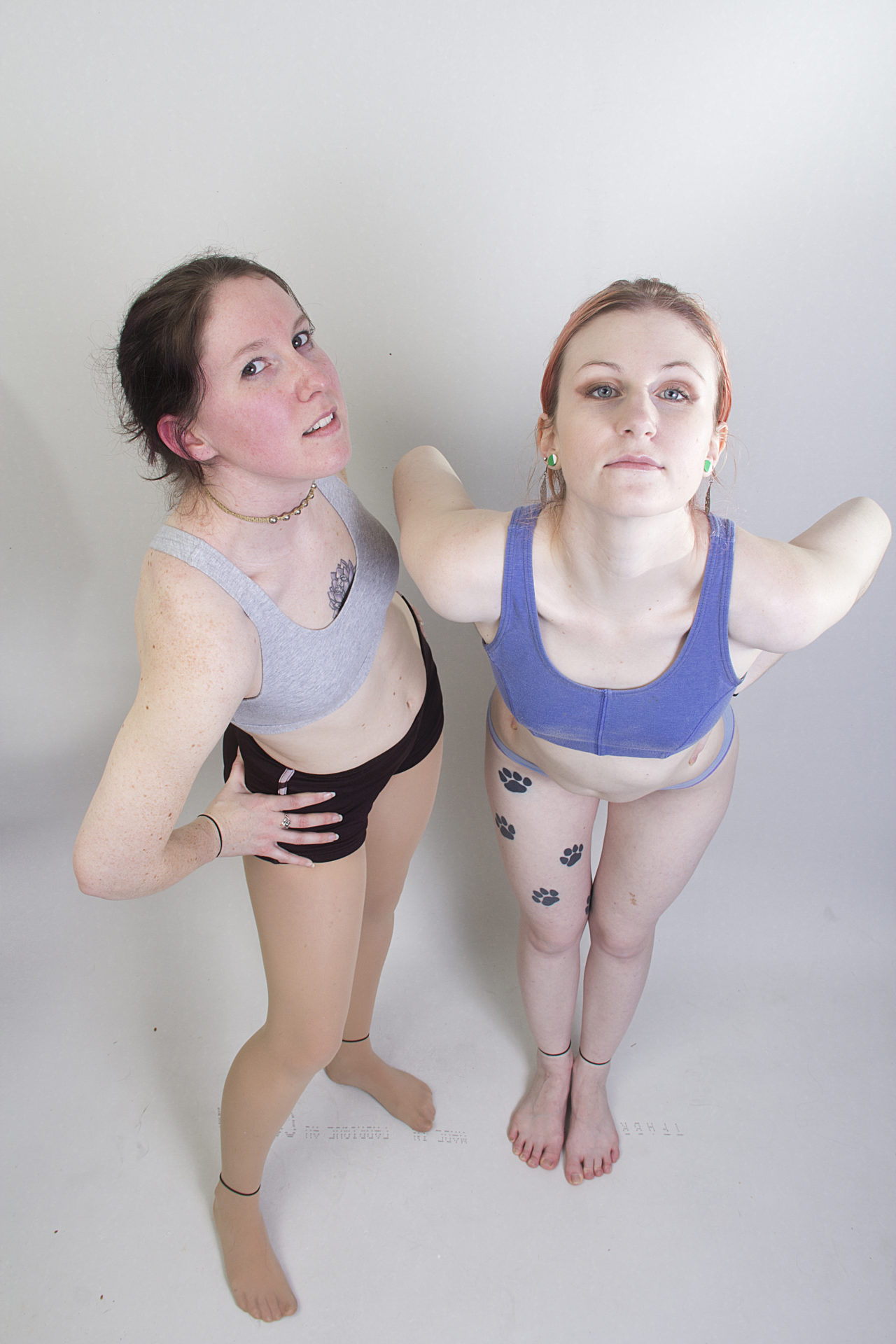 Two thin, white, female models wearing sports bras and short shorts, from a high angle perspective, standing with hands on their hips and leaned slightly forward as they both glare directly into the camera.