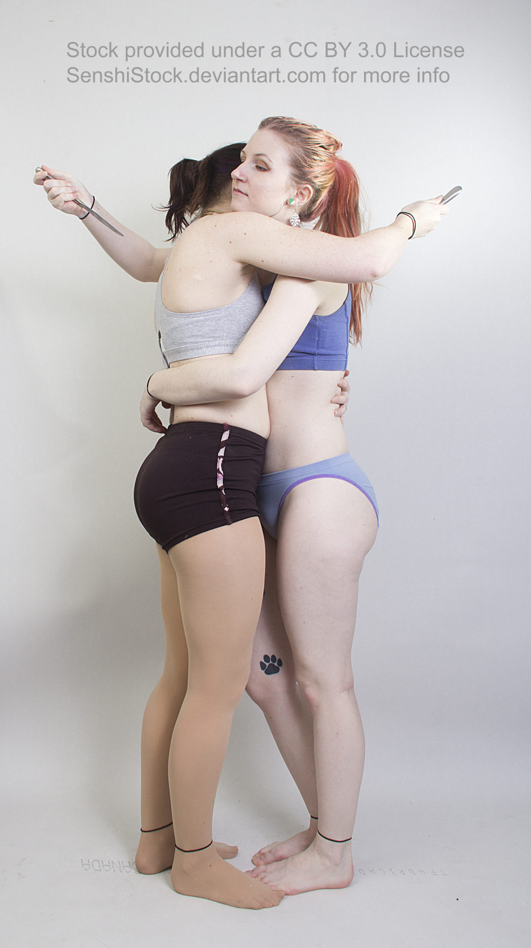 Two thin, white, female models wearing sports bras and short shorts, standing hugging each other, both holding a small knife about to stab each other in the back.