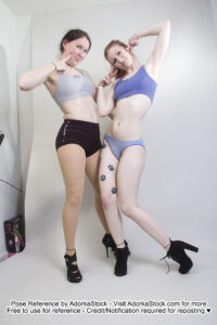 Two thin, white, female models wearing sports bras and short shorts and black high heels. Both are standing next to each other, smiling at the camera. One is poking a finger at her cheek. The other is giving peace signs by her cheeks, with one leg bent and relaxed.