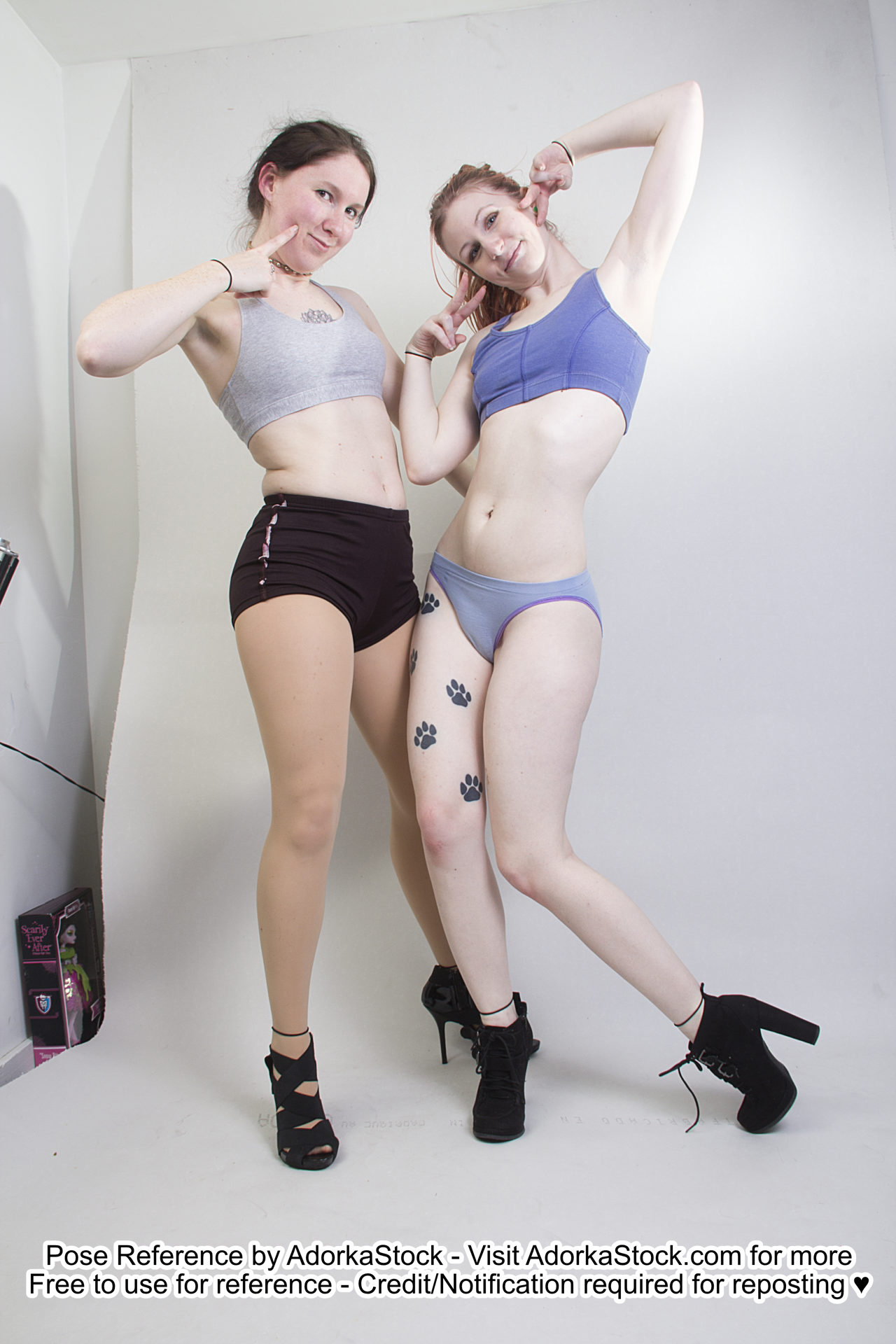 Two thin, white, female models wearing sports bras and short shorts and black high heels. Both are standing next to each other, smiling at the camera. One is poking a finger at her cheek. The other is giving peace signs by her cheeks, with one leg bent and relaxed.