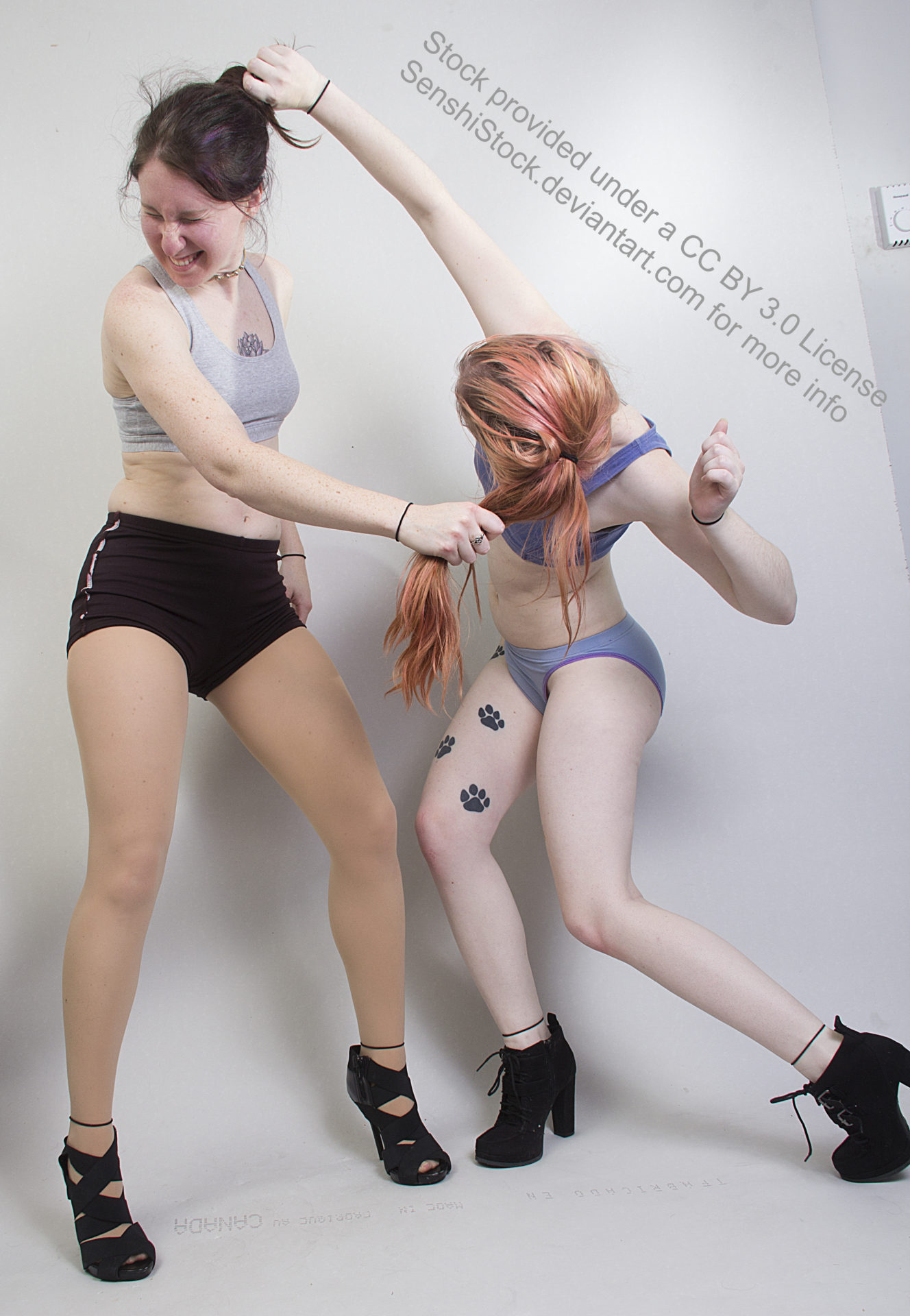 Two thin, white, female models wearing sports bras and short shorts with black high heels in a fight. They're both pulling at each other's hair, one standing upright, making a pained face, the other crouched down as she's reaching up to grasp the other's hair.