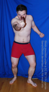 A muscular white man wearing a pair of red boxers is standing with his knees slightly bent and legs spread apart. One arm is pulled back and in a fist, the other is out at the camera while holding a knife. His head is down and he's glaring down the camera.