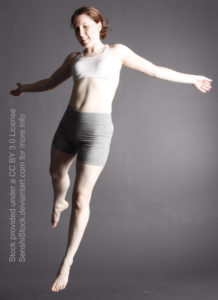 White woman jumping in the air, arms outstretched, wearing a white top and grey skin tight pants.