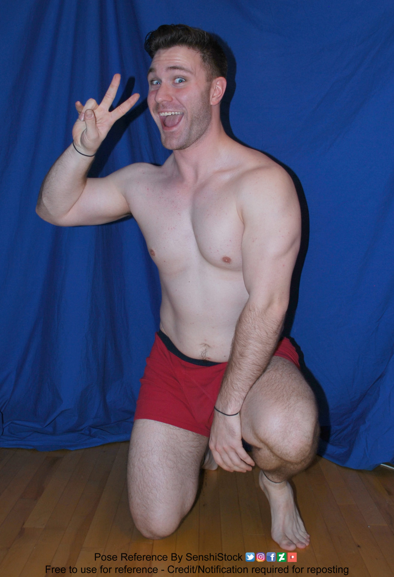 A muscular white man wearing a pair of red boxers is kneeling on one knee with his arm resting on the other, his other arm is raised up to his head and has two fingers raised for a peace sign. He has a massive grin on his face.