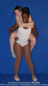 A Black woman wearing a white leotard and pink ballerina slippers holding a white woman on her shoulders in a piggyback ride.