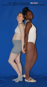 Two women, one white and the other black, wearing skin tight clothing, standing back to back, holding hands, and looking at the camera.