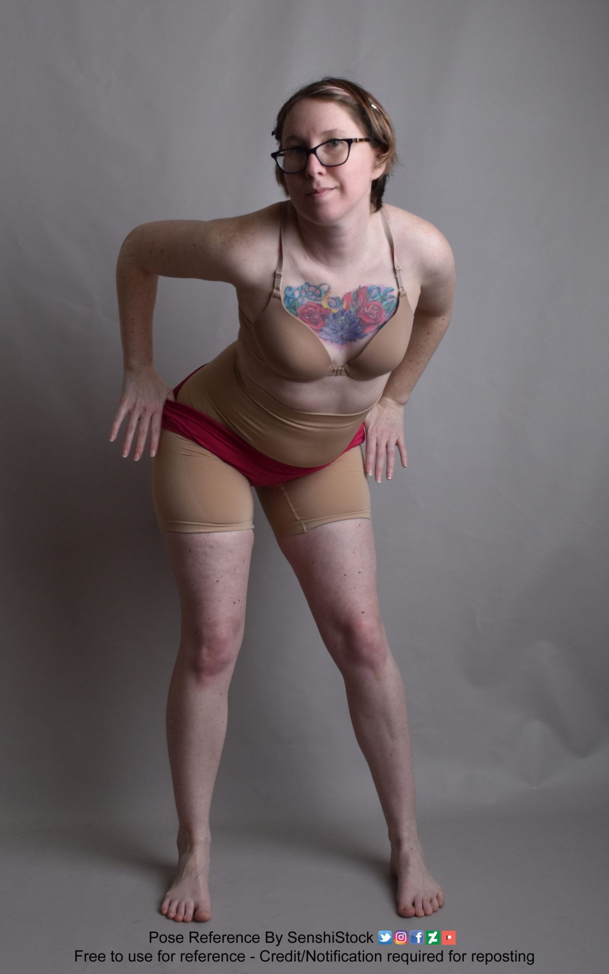 Thin white woman wearing a form-fitting beige bra and waist high bottoms and glasses, standing leaning forward toward the camera with hands pulling at an undergarment as she smiles at the camera.