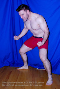 A muscular white man wearing a pair of red boxers is squatting slightly, arms tensed, bent at the elbows, hands in a fist, as his face is in an angry grimace expression.