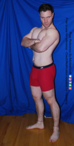 A muscular white man wearing a pair of red boxers is standing 3/4 with his arms crossed over his chest. His head is turned toward the camera as he's glaring.