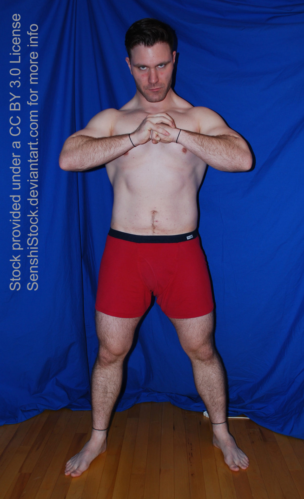 A muscular white man wearing a pair of red boxers is standing facing the camera, legs spread apart, arms raised as he's smacked a fist into the palm of his other hand. He's glaring down the camera.