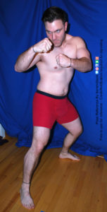 A muscular white man wearing a pair of red boxers is standing with his legs spread apart in a stable stance, knees slightly bent, with both arms raised in fists, as he's glaring past the camera.