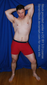 A muscular white man wearing a pair of red boxers is standing with his legs spread apart, one knee slightly bent, both arms are raised above and behind his head as he's giving a sultry glare at the camera.