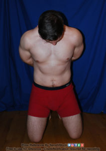 A muscular white man wearing a pair of red boxers is on his knees, arms behind is back, head down.